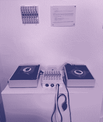 an installation of two turntables on a platform, with a box connected to headphones, an inflation bulb, and a mouthpiece. instructions are mounted on the wall